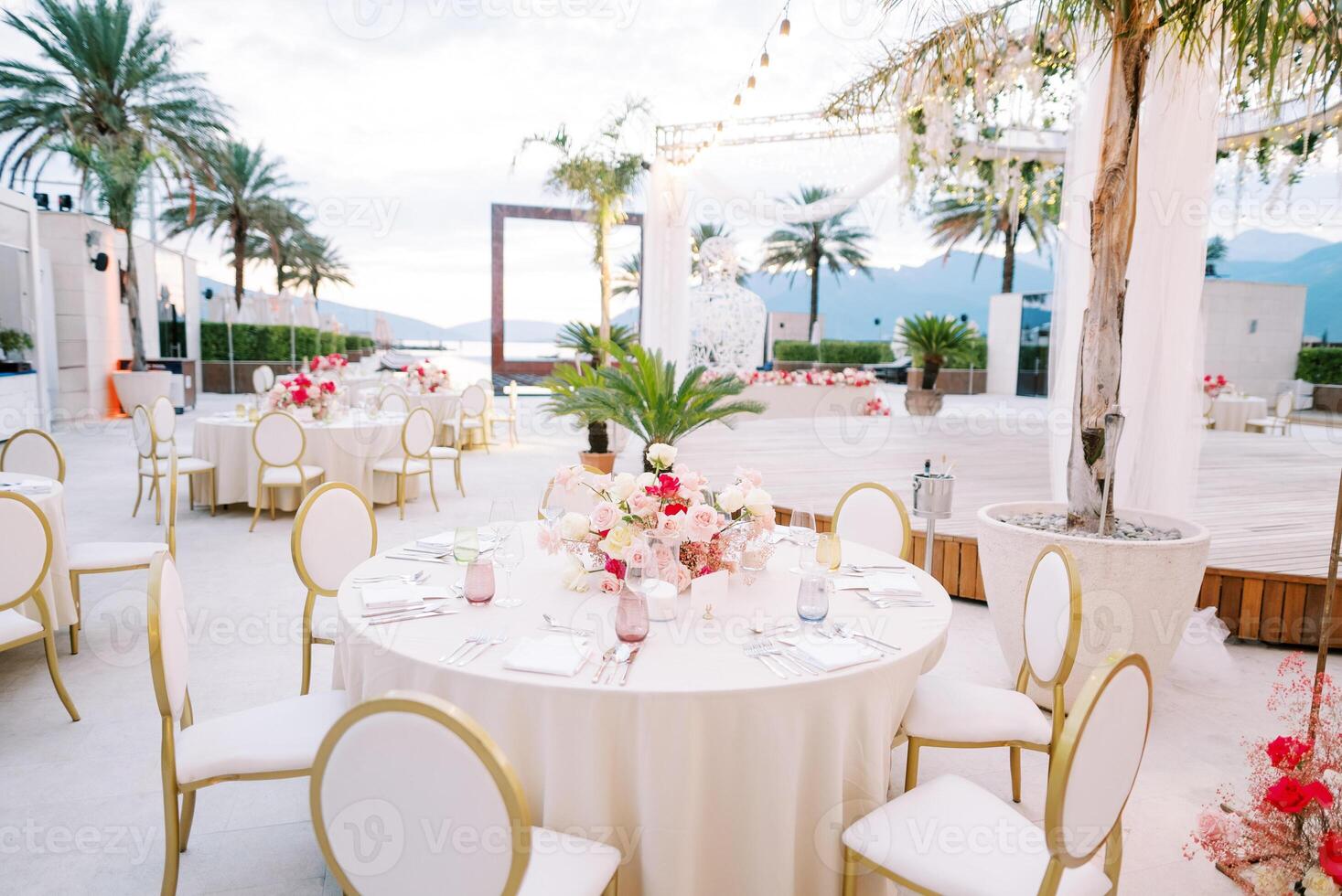 Festive tables with bouquets of flowers and white chairs stand near a stage decorated with garlands of light bulbs photo