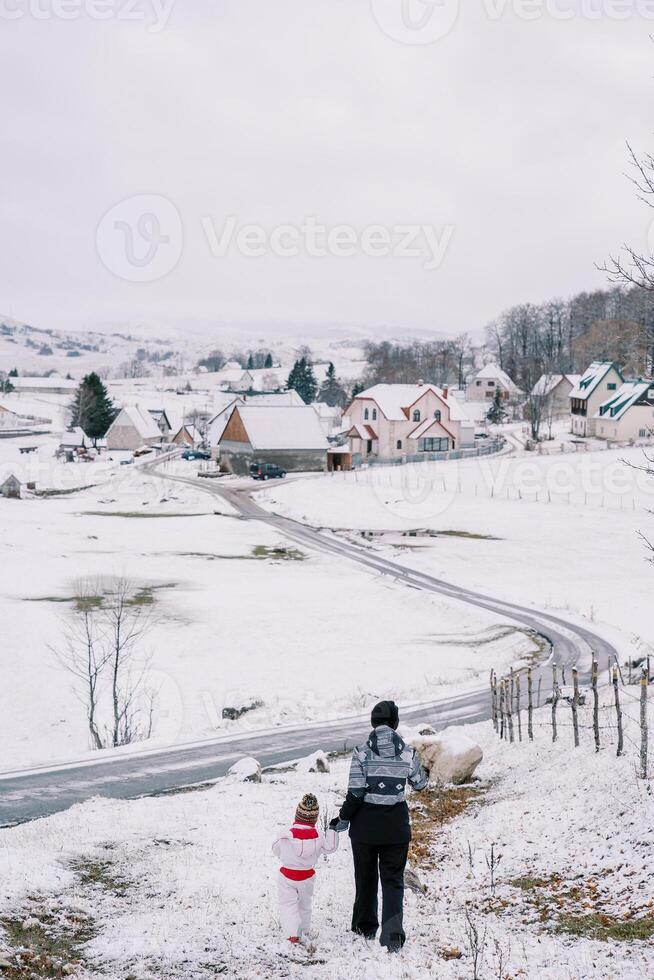 madre y un pequeño niña caminar abajo un Nevado colina participación manos hacia un pueblo la carretera. espalda ver foto