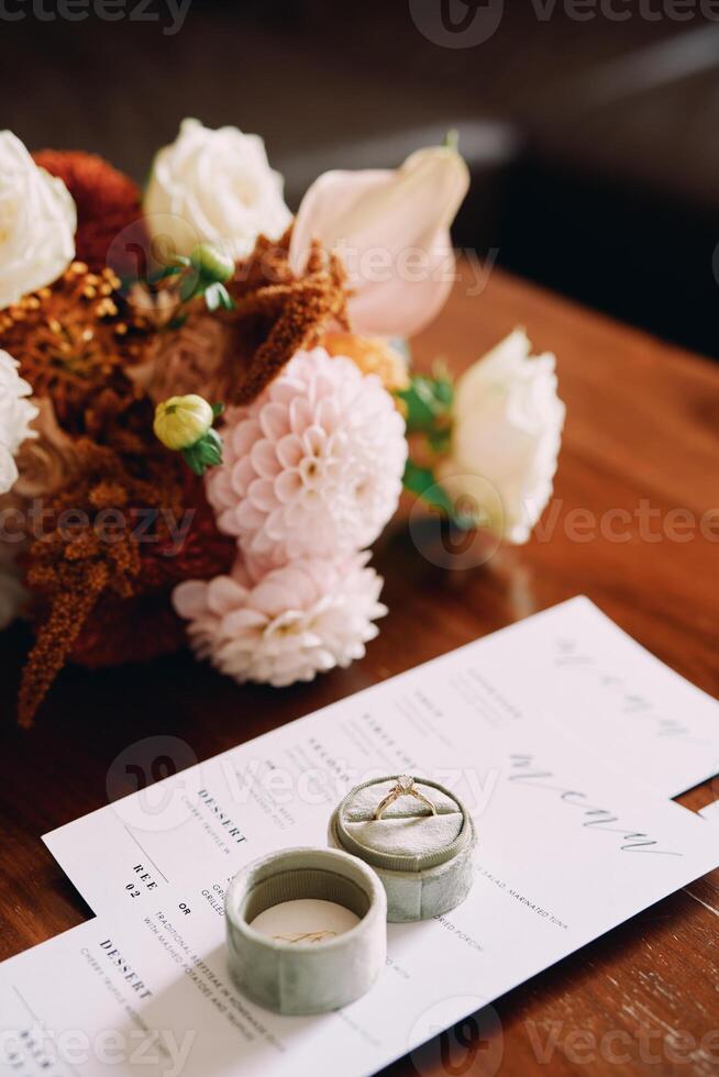 Engagement ring in a box stands on a festive menu on a wooden table near a bouquet photo