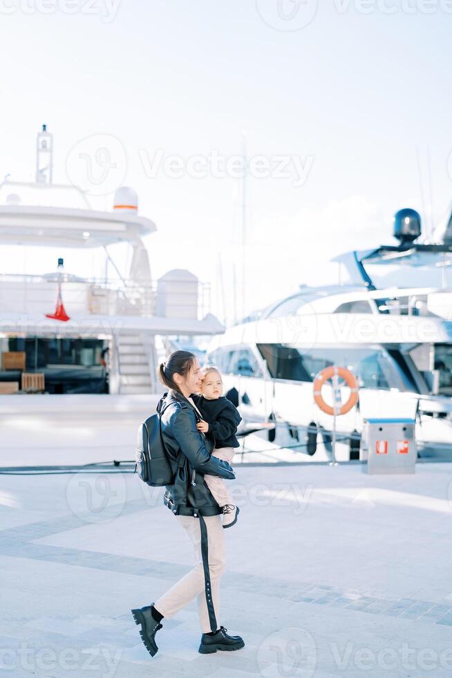 Mother with a little girl in her arms walks along the promenade past moored yachts. Side view photo