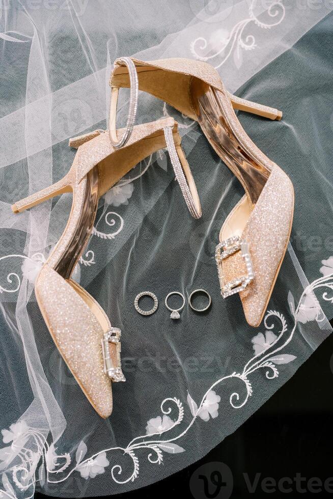 Wedding rings lie on an embroidered veil near the bride golden high-heeled shoes photo