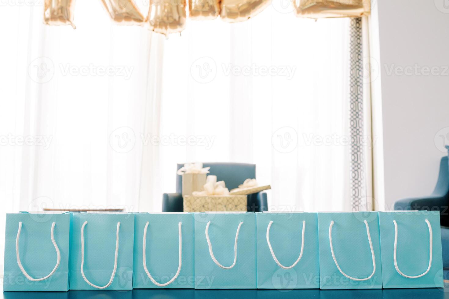 Row of blue gift bags with white handles stands on a table in the room photo