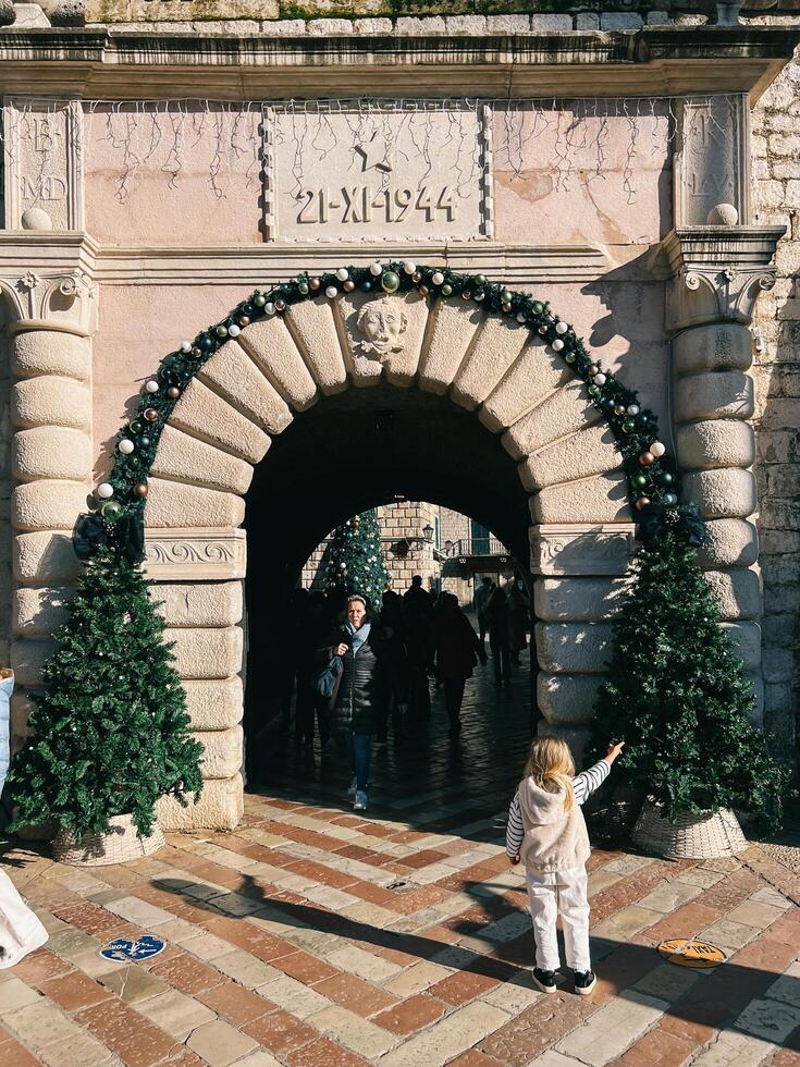 Kotor, Montenegro - 25 december 2022. Little girl stands in front of an ancient arch and points to a decorated Christmas tree photo