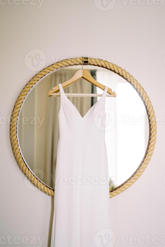 Wedding dress hangs on a hanger on a round mirror in a rope frame on the wall in the room photo