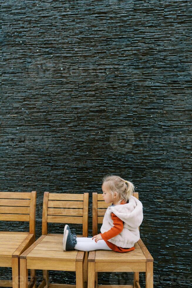Little girl sits on a row of wooden chairs against the wall and looks at her shoes. Side view photo