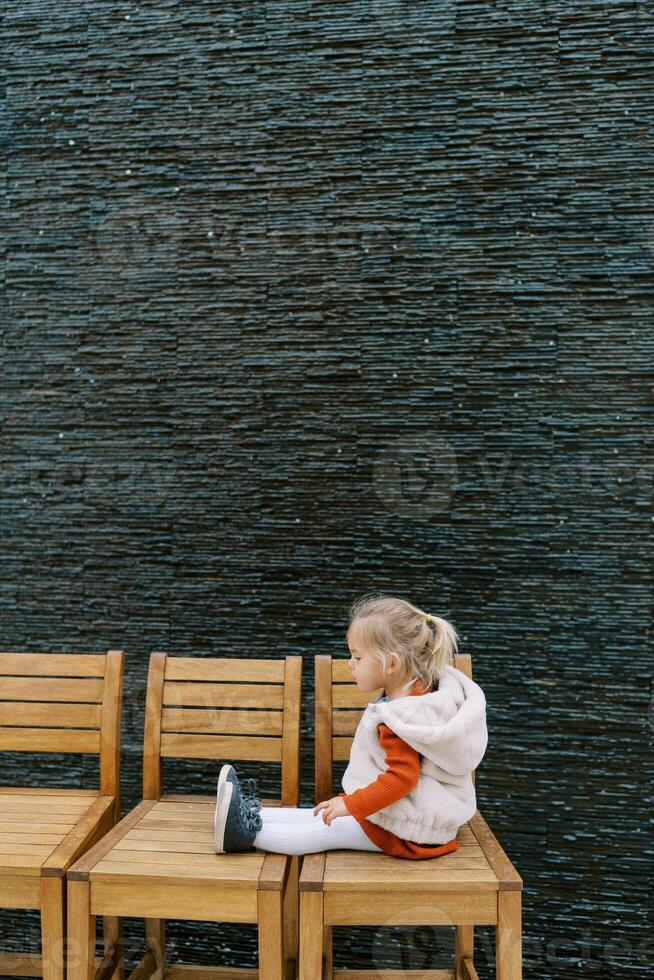 Little girl sits on a row of wooden chairs against the wall and looks at her shoes. Side view photo