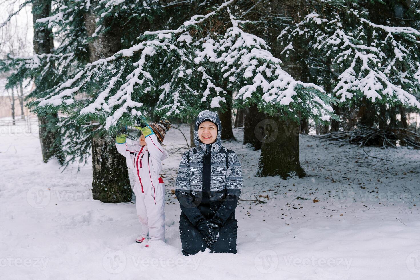 Smiling mother in a ski suit sits on her knees in the snow near a small child standing near a snow-covered pine tree photo