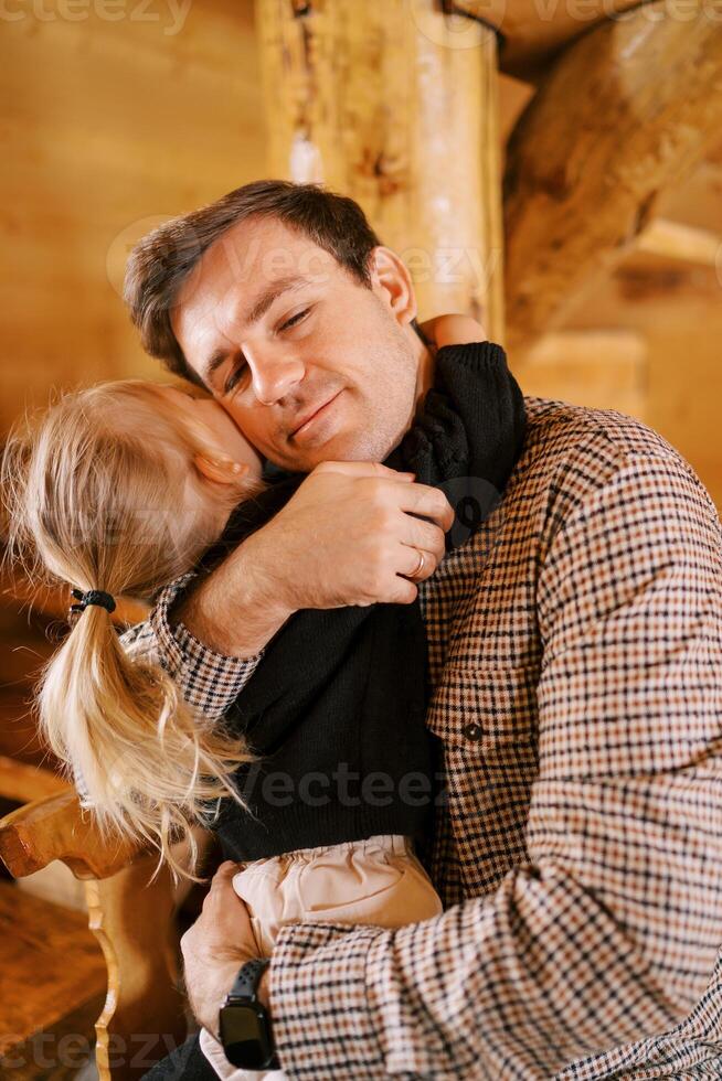 Dad hugs a little girl sitting on his lap on a wooden chair near the stairs photo