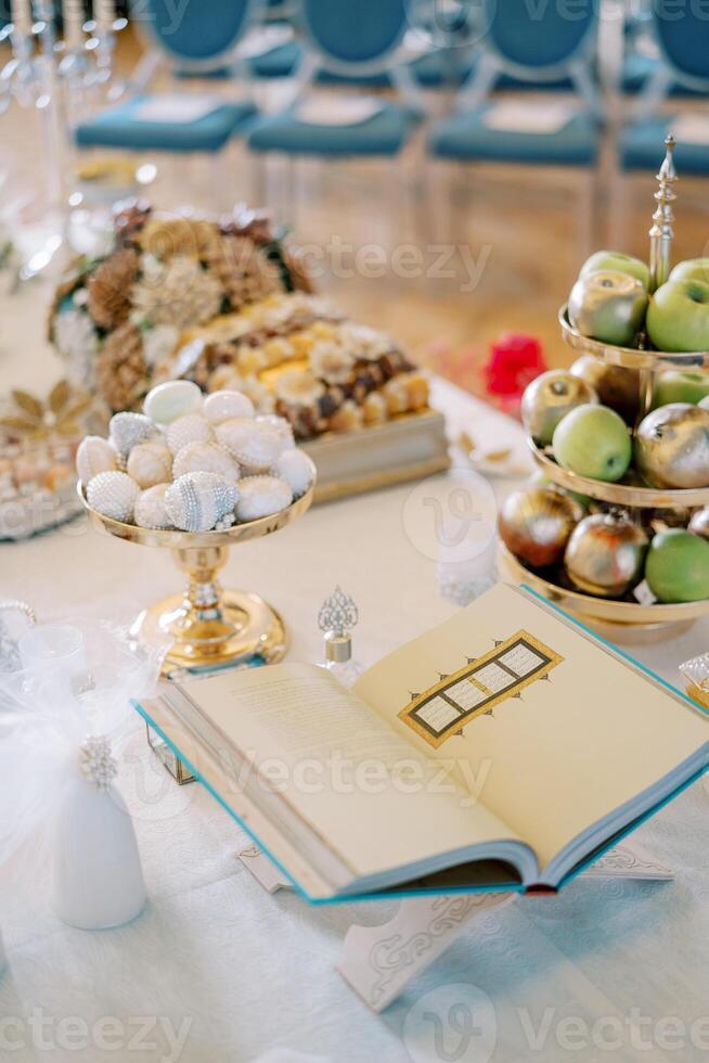Open holy book on a stand on a table next to fruit in vases, pastries, sugar cones and nuts. Tradition of Sofreh Aghd photo