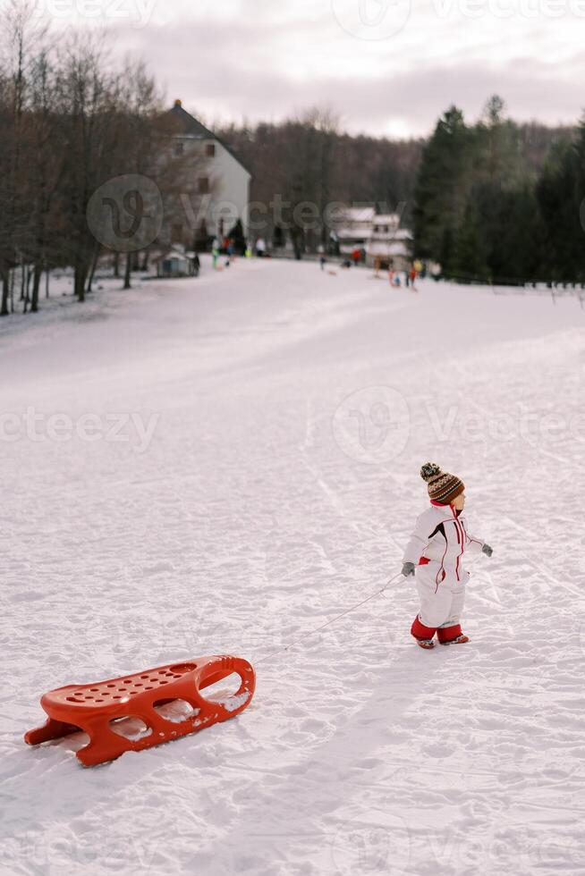 Little girl is walking along a snowy hill, dragging a sled on a rope behind her. Side view photo