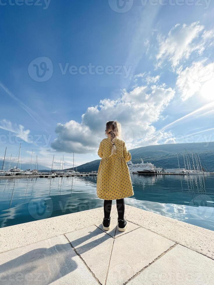 Little girl stands on the pier and looks at the marina with yachts on the background of the mountains. Back view photo