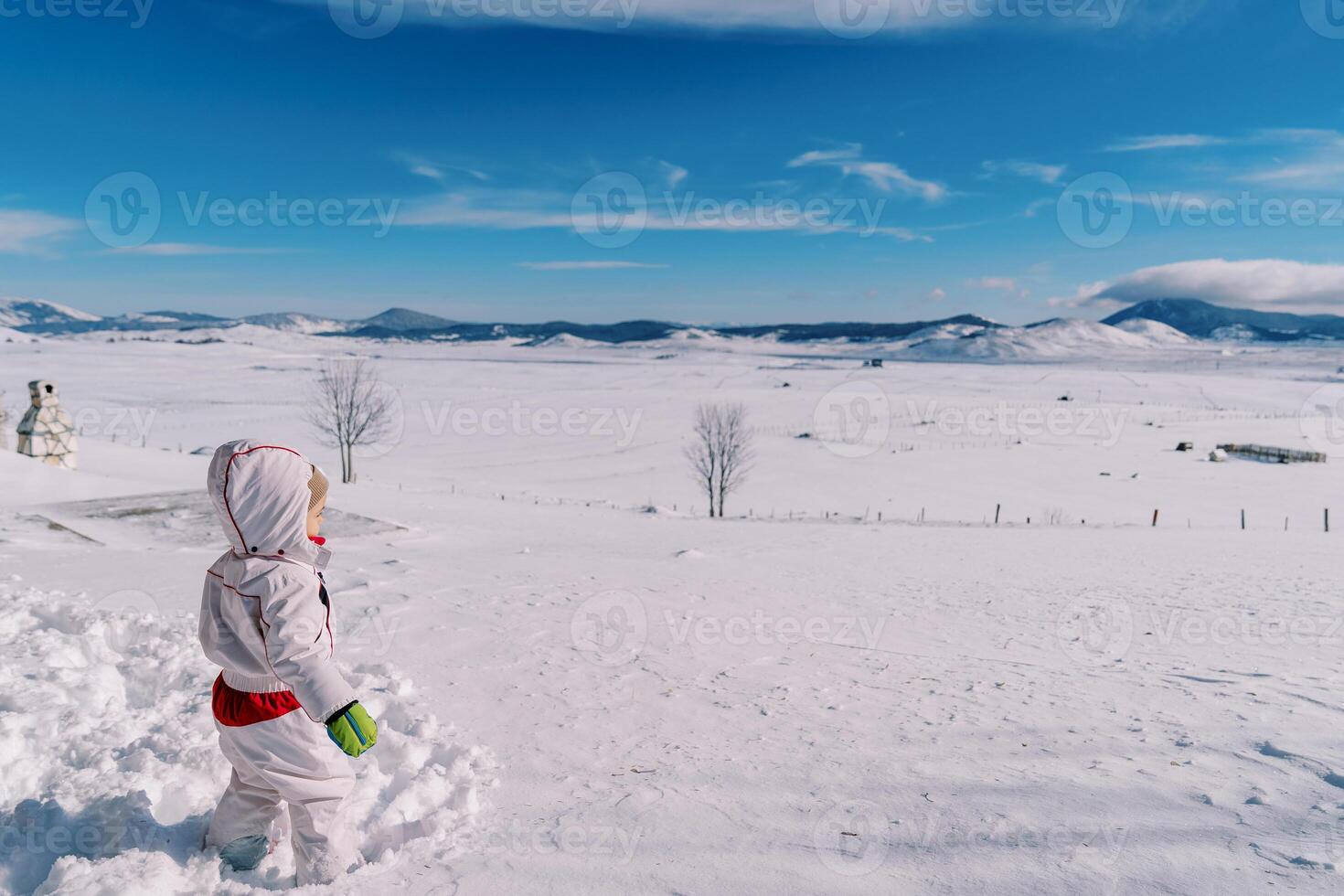 Little girl stands in a deep snowdrift in a mountain valley and looks into the distance. Side view photo