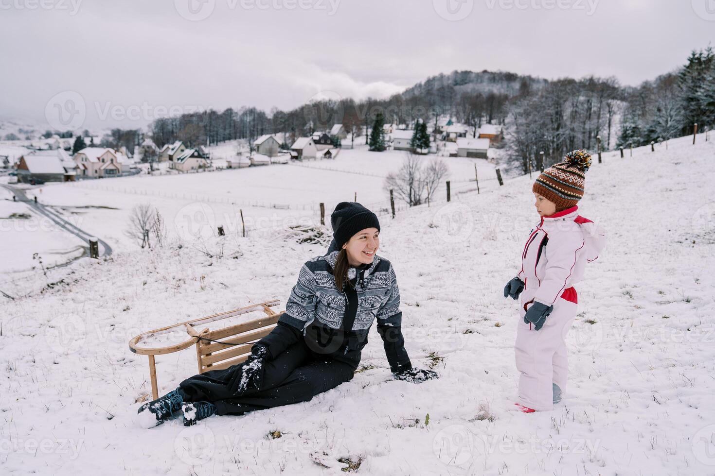 Little girl stands on a snowy hill next to a sitting smiling mother and a sled turned on its side photo