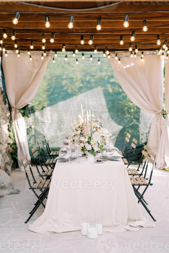 Laid table with bouquets of flowers stands on the terrace of an old house under a wooden canopy with curtains photo