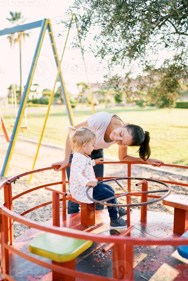 Mom leaned over to a little girl sitting on a carousel on the playground photo