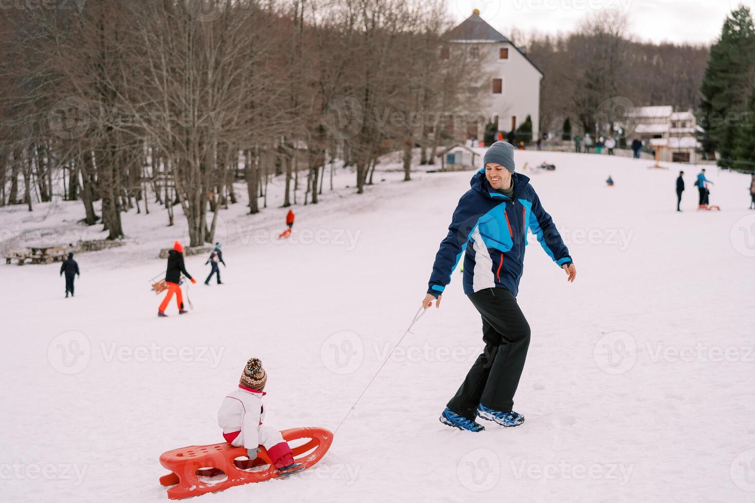 Smiling dad climbs a hill with a small child in a sled, looking back. Side view photo