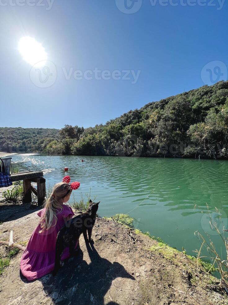 Little girl with Minnie Mouse ears on her head sits on the river bank next to a cat photo