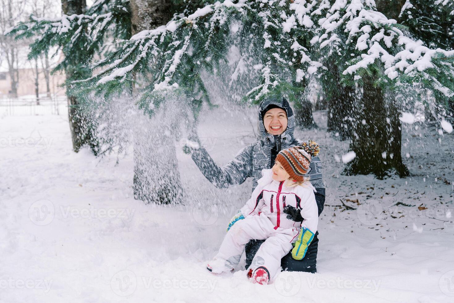 sonriente mamá con un pequeño niña se sienta en su rodillas en el nieve debajo nevada desde pino ramas foto