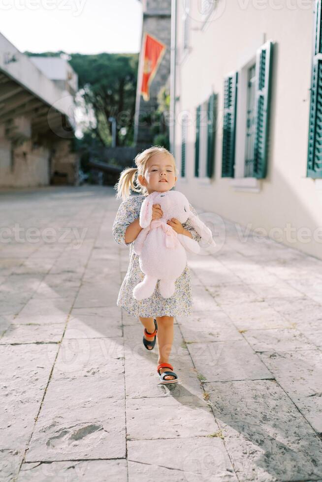 Little girl walks hugging a pink toy rabbit through the courtyard of an old castle photo