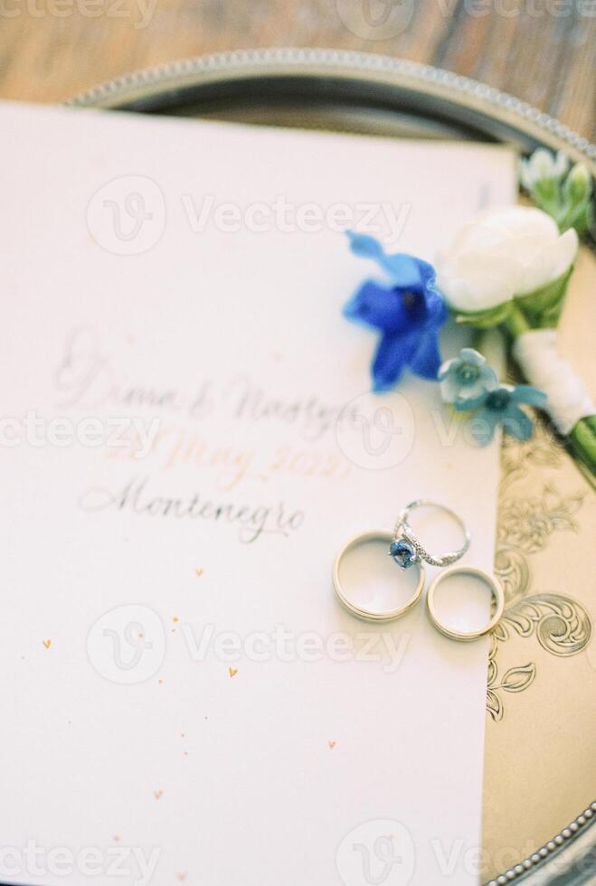 Wedding rings lie on an invitation next to a boutonniere on a tray photo