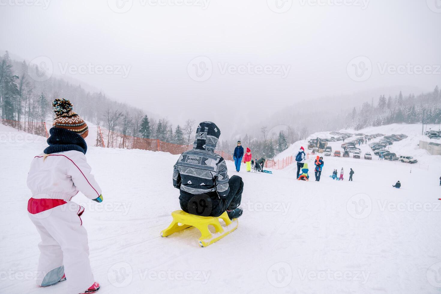 Small child looks at his mother riding a sled down a hill. Back view photo