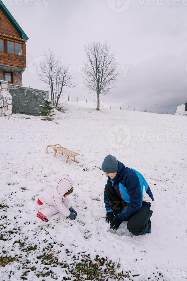 Dad and a little girl are squatting on a hill near a sled and making snowballs photo