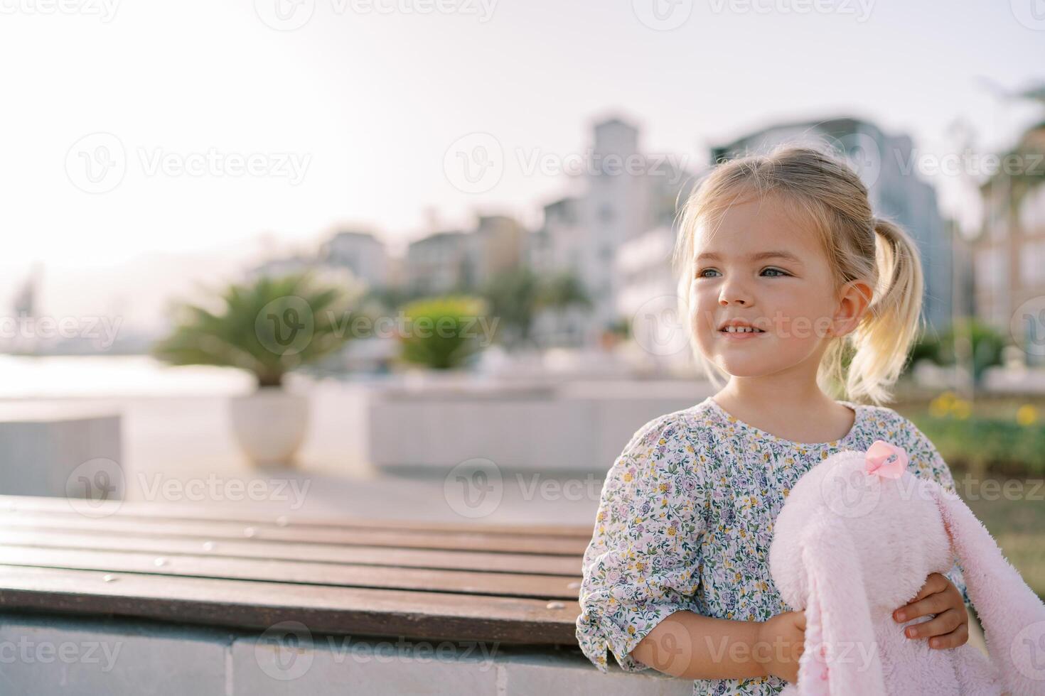 Little smiling girl with a pink soft toy hare stands near a wooden table on the embankment and looks away photo