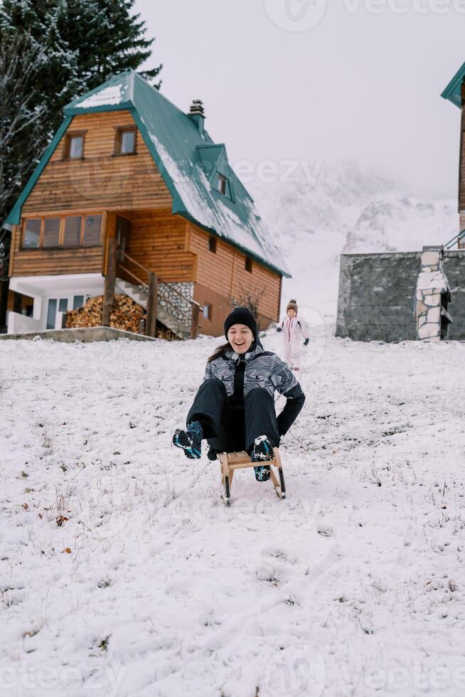 Little girl stands near the house and looks at her laughing mother riding down the hill on a sled photo