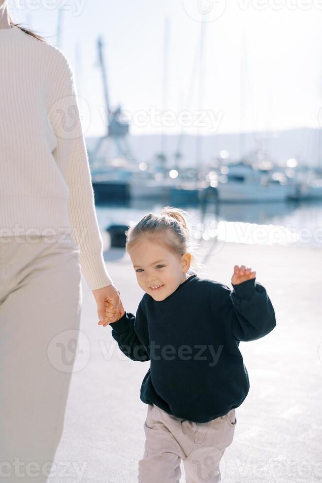 Little smiling girl walks along the pier holding her mother hand and waving. Cropped photo