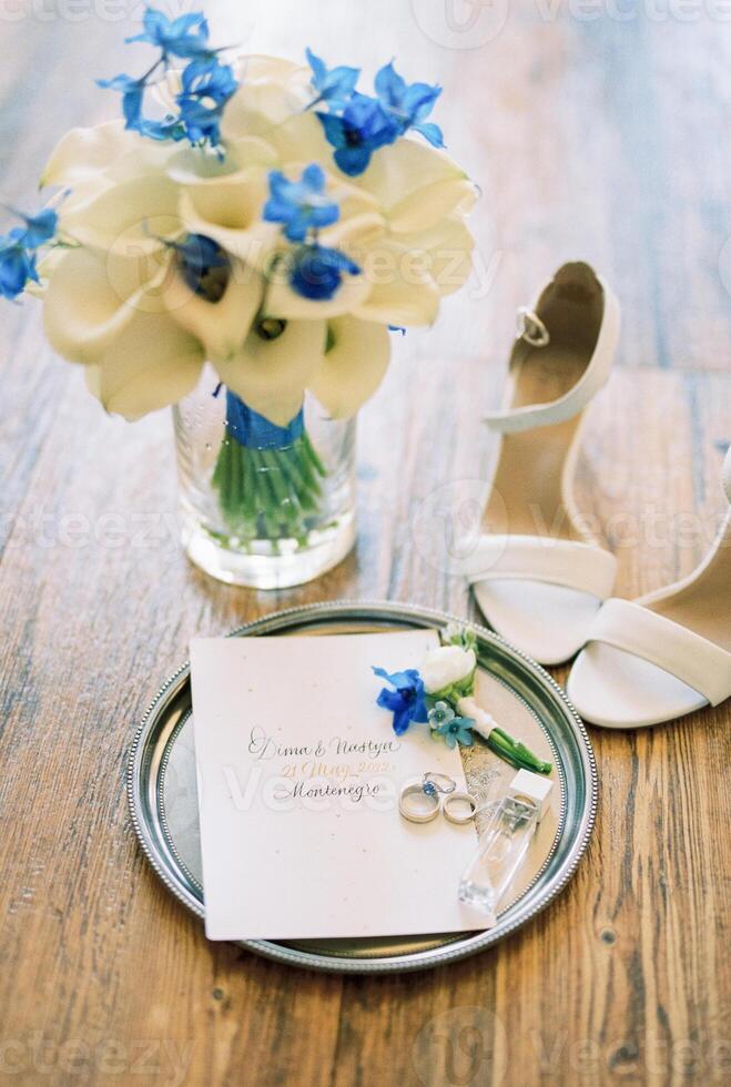 Wedding rings lie on a round tray next to the invitation near a wedding bouquet of flowers in a vase photo
