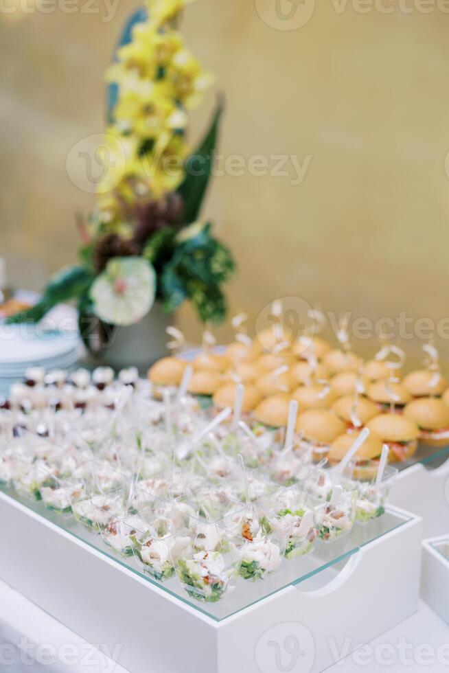 Portioned salads in cups with spoons on a tray next to mini-burgers are on the table photo