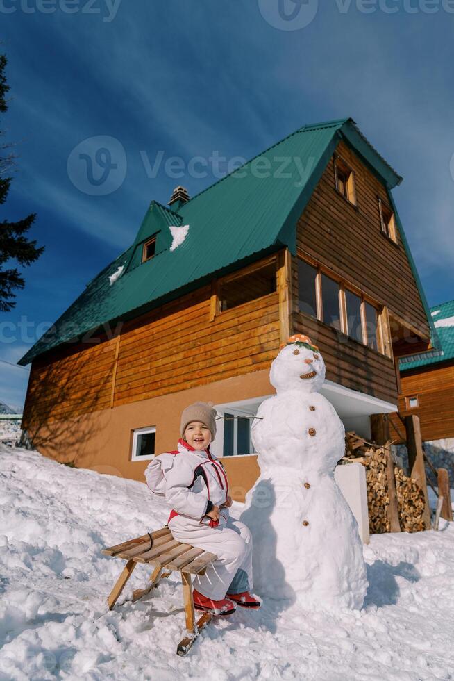 Little girl sits sideways, opening her mouth on a sleigh near a snowman in the yard photo