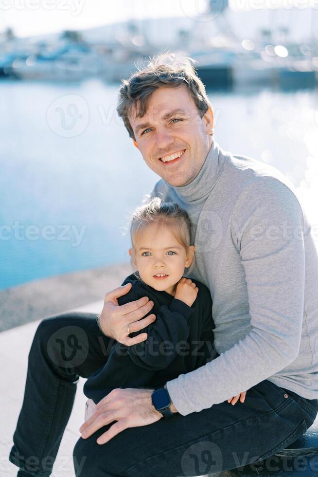 Smiling dad hugs little girl leaning on his knee while sitting on a pillar on the pier photo