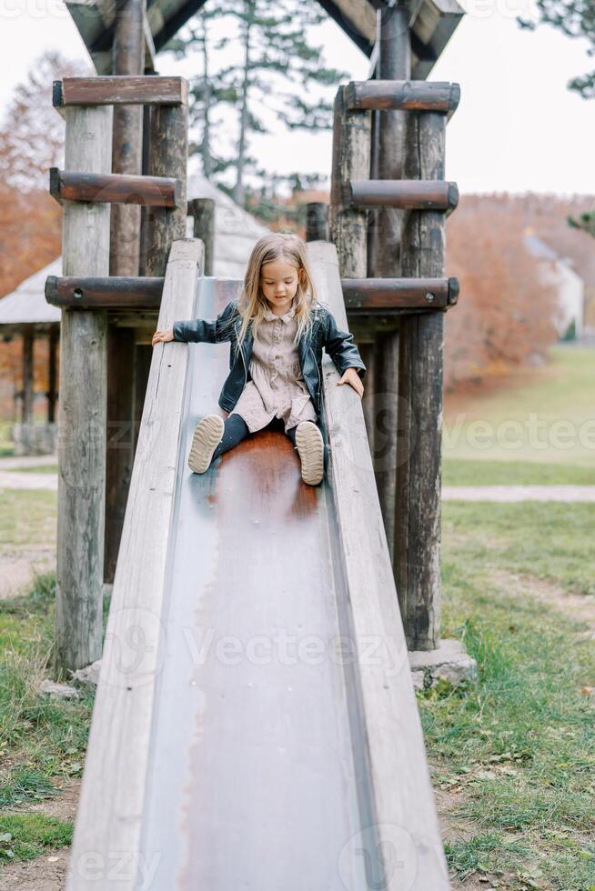 Little girl slides down a slide on a wooden playground holding the handrails photo