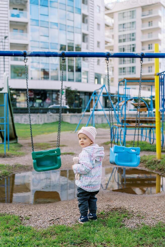 Little girl stands near the swing in front of a puddle, turning around. Back view photo