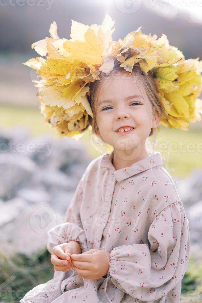 Little smiling girl in a wreath of autumn leaves sits on a stone on the lawn photo