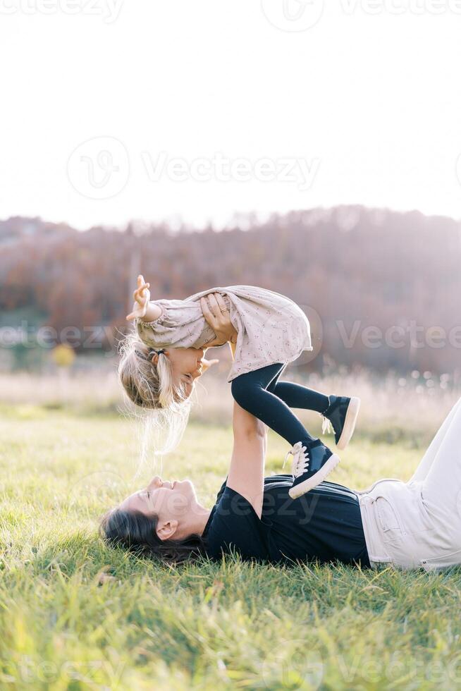 Smiling mother lifting little girl with arms outstretched to sides while lying on green lawn photo