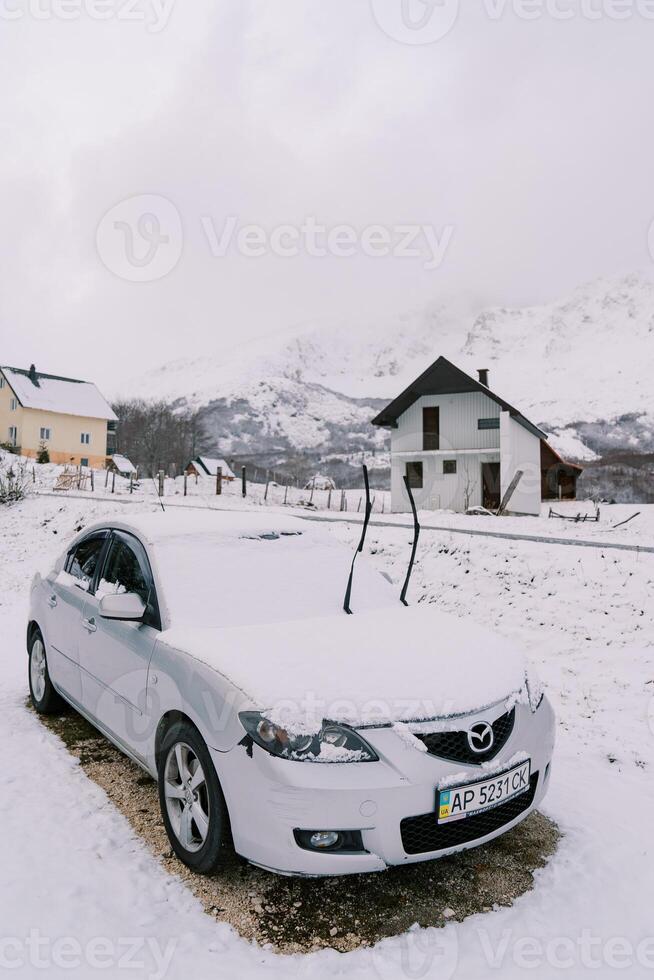 Snow-covered car with raised windshield wipers stands near a cottage photo