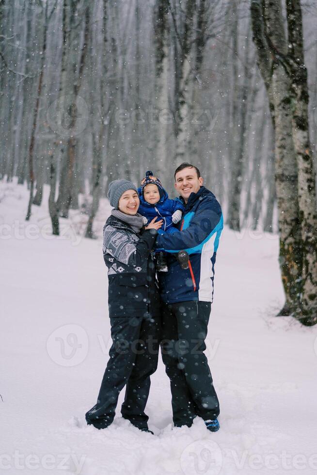 Smiling parents with a small child in their arms stand under snowfall in the forest photo