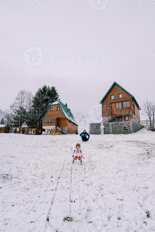 papá sentadillas cerca el casa, acecho un pequeño niña trineo abajo un Nevado colina foto