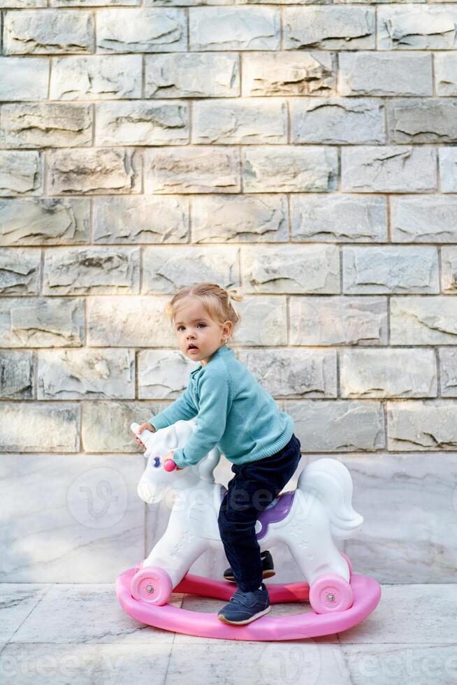 Little girl sitting on a toy unicorn rocking chair near a stone house photo