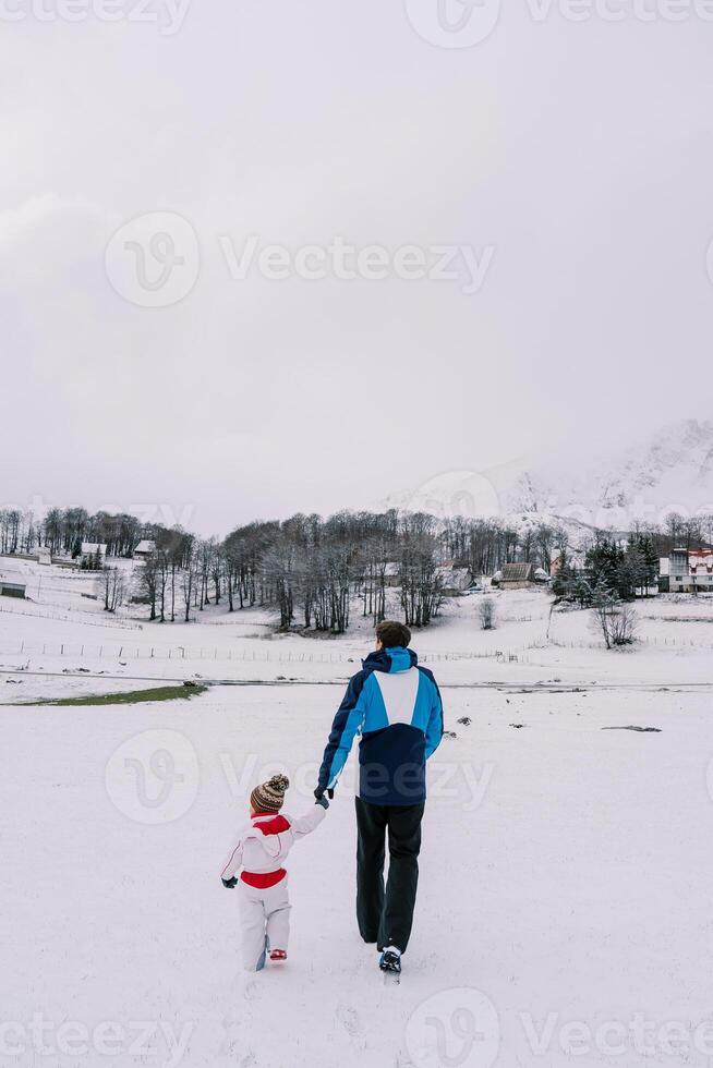 Dad and small child walk holding hands through a snowy village pasture. Back view photo