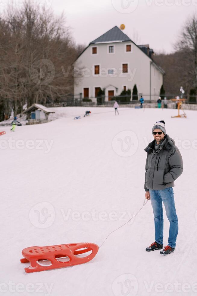 Smiling man with a sled on a rope stands on a snowy hill photo