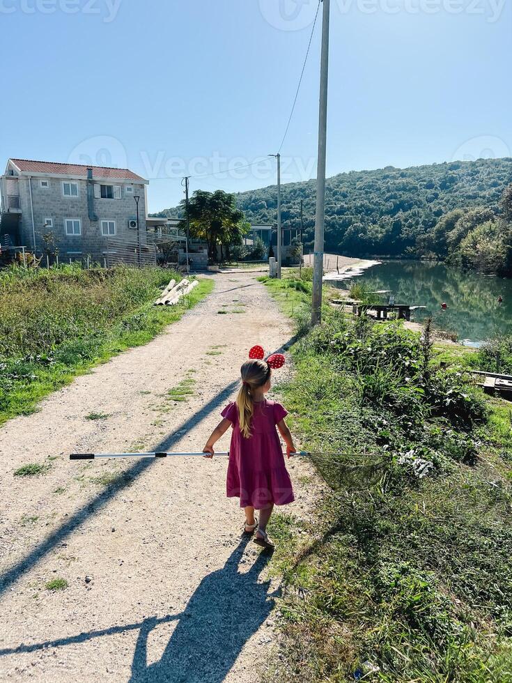 Little girl with Minnie Mouse ears on her head and a landing net in her hands walks along the river bank photo