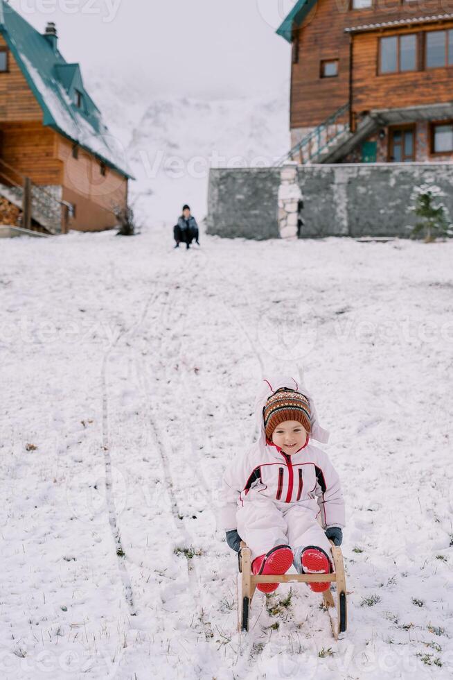 pequeño sonriente niña diapositivas abajo un Nevado colina en un trineo en contra el fondo de su mamá sentado cerca el casa foto