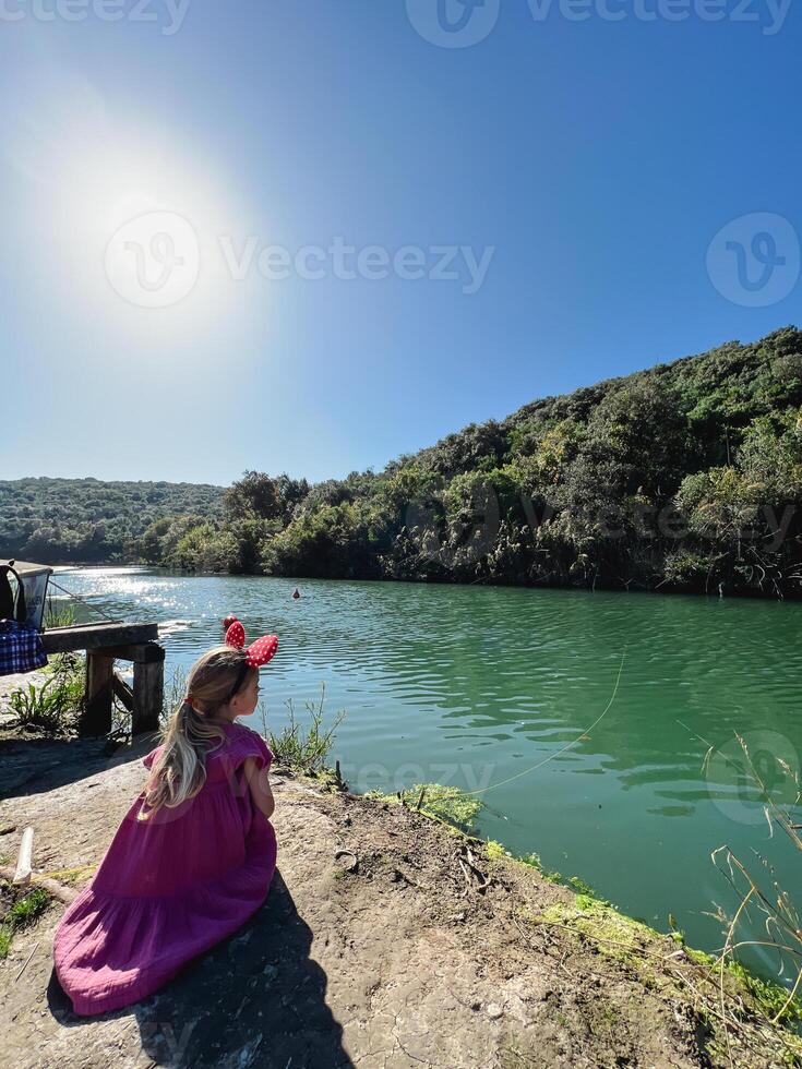 Little girl with Minnie Mouse ears on her head sits on the river bank photo