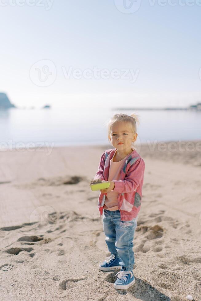 pequeño niña soportes en el playa con un juguete pala lleno de arena foto