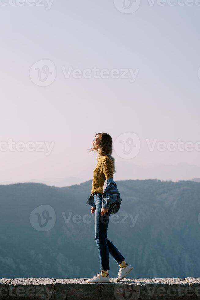 Young woman in sunglasses stands on a stone curb high in the mountains. Side view photo