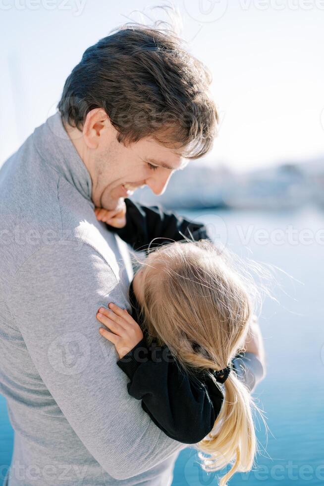 sonriente papá sostiene un pequeño niña abrazando su hombro mientras en pie por el mar foto