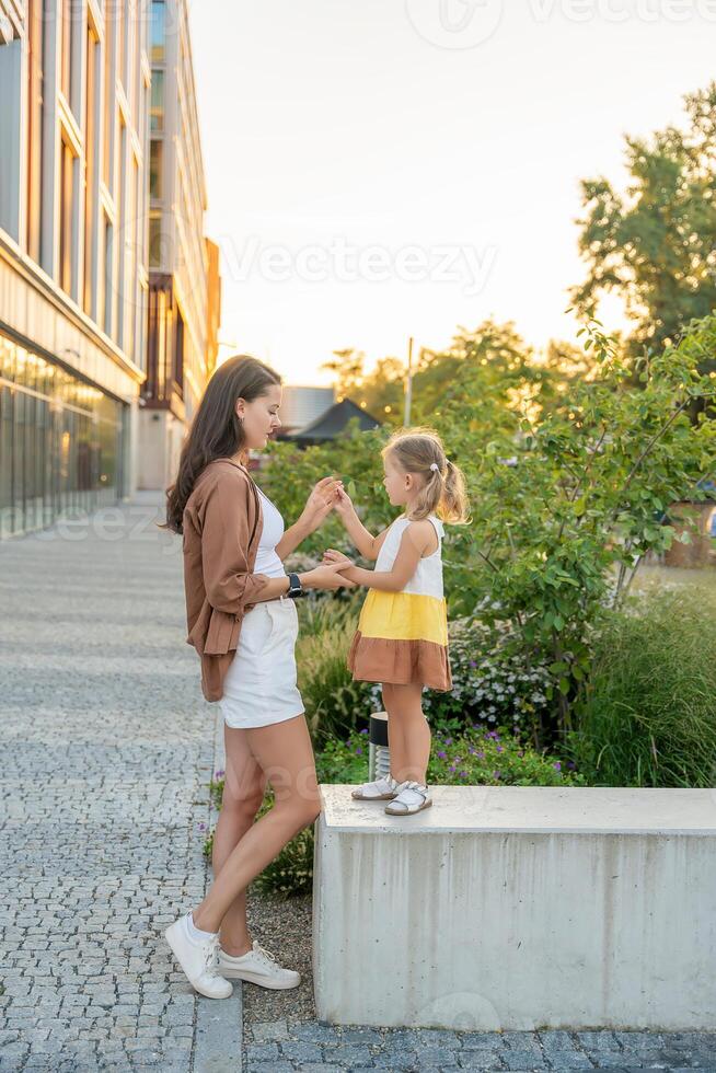 mamá y su pequeño hija jugando en ciudad calle en puesta de sol tiempo. alto calidad foto